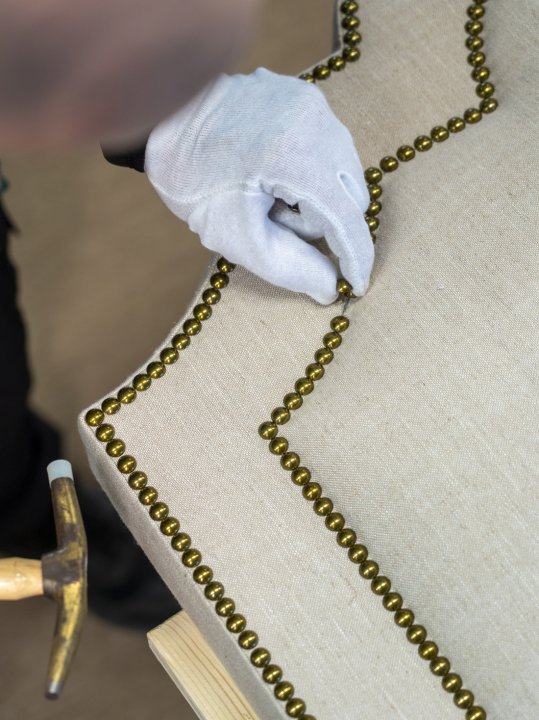Behandschuhte Hand setzt Messingnägel in ein gepolstertes Bettkopfteil in der Elite-Manufaktur.
