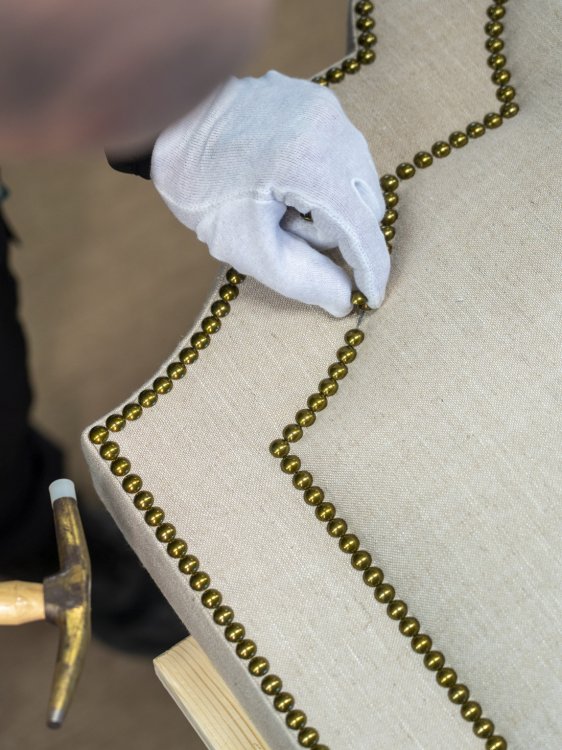 Behandschuhte Hand setzt Messingnägel in ein gepolstertes Bettkopfteil in der Elite-Manufaktur.