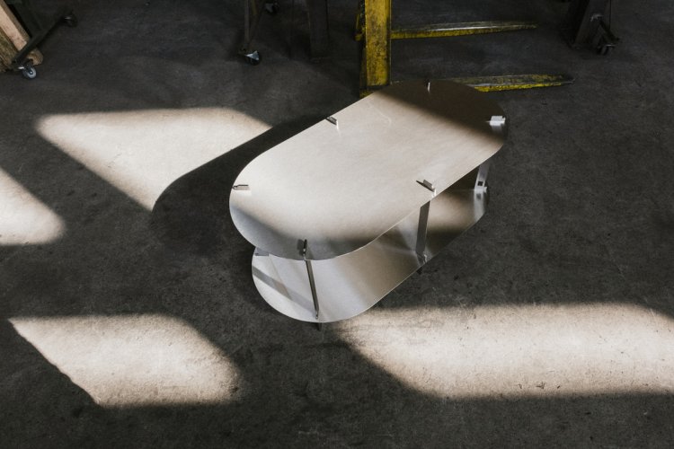 Ein ovaler, zweistöckiger Tisch aus geschliffenem Chromstahl steht auf einem Betonboden, auf den Licht und Schatten fallen.