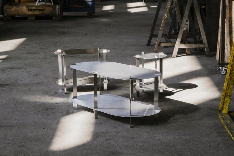 Drei minimalistische Tische aus Metall in unterschiedlichen Formen und Grössen stehen auf einem Betonboden in einer Werkstatt, beleuchtet von einfallendem Tageslicht.