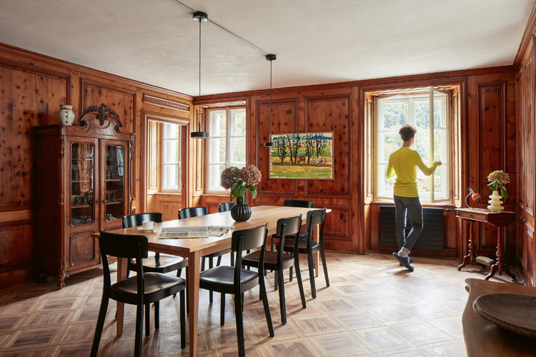 Ein Raum mit Holz ausgekleidet, ein Esstisch, ein Mann in einem gelben Pullover am Fenster
