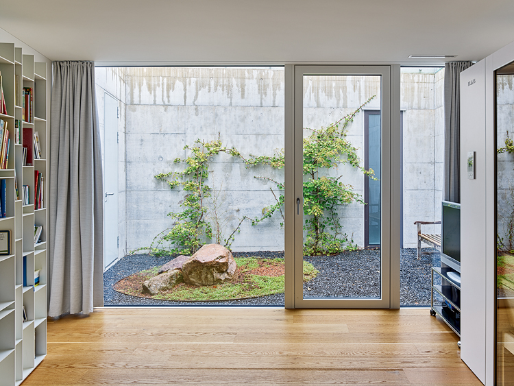 Bibliothek in einem Einfamilienhaus, diese sieht man nur seitlich angeschnitten, der Blick fällt durch eine Fensterfront ins Atrium, wo inmitten des Kiesbodens eine grüne Oase ist.