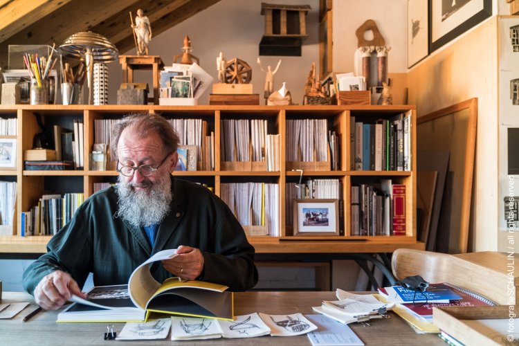 Aufnahme von Michele de Lucchi sitzend und blätternd in einem Buch vor einer Bücherwand in seinem Atelier.