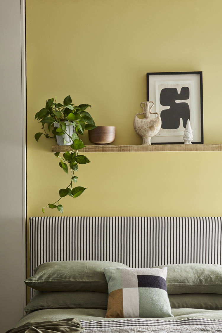 Graues Bett mit grüner Bettwäsche vor gelber Wand.