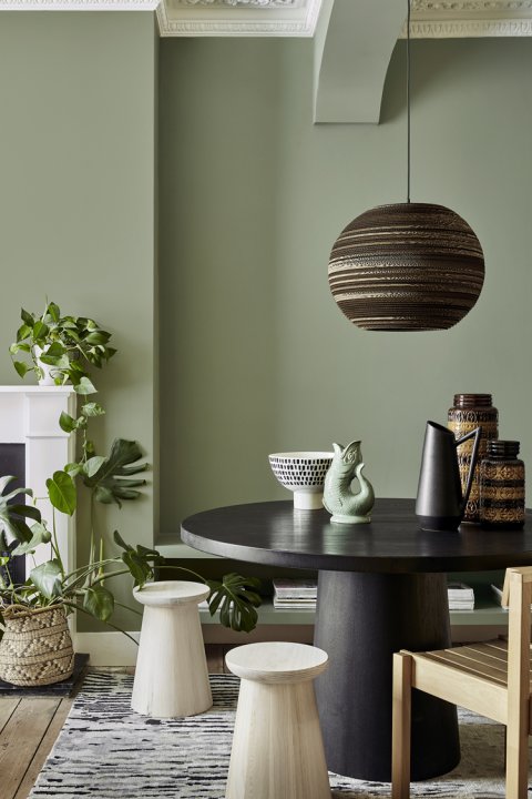 Wohnzimmer mit grüner Wand und schwarzen rundem Tisch davor umzingelt von hellen Hockern und einem Holzstuhl.
