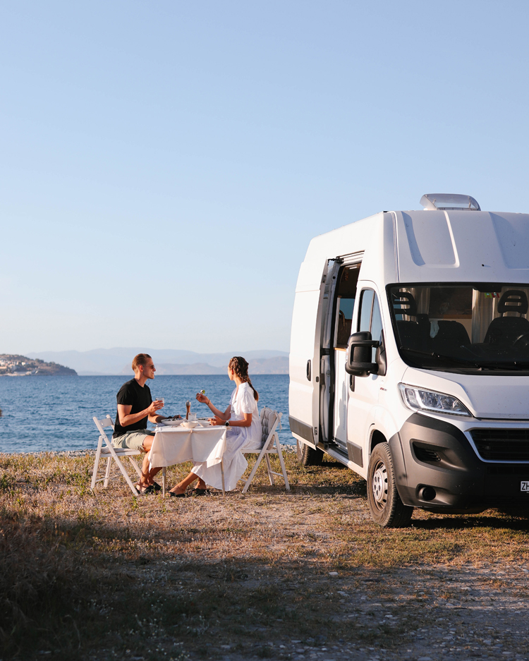 Paar sitzt neben weissem Camper-Van in der Sonne.