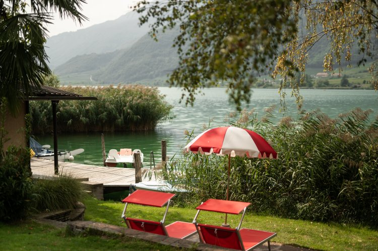 Wiese direkt am See mit roten Liegestühlen und weiss-roten Sonnenschirmen hinter Schilf.