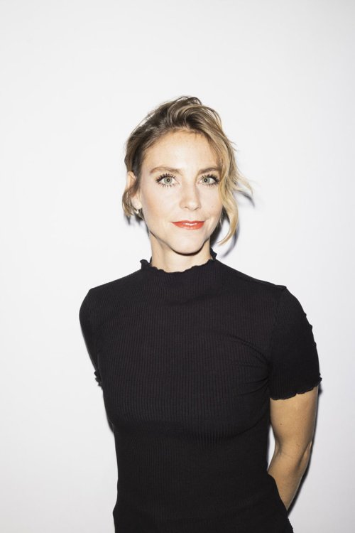 Porträt von Designerin Simone Züger mit hellem Teint, blondem Haar und schwarzem Kurzarm-Oberteil, die vor einer weissen Wand in die Kamera blickt.