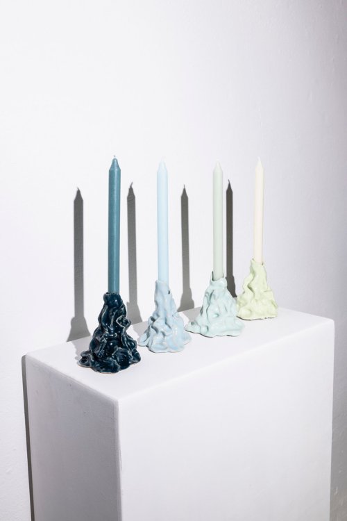 Vier farblich abgestufte Kerzen in handgeformten, organisch wirkenden Kerzenständern aus glasiertem Ton, auf einem weissen Sockel vor einer weissen Wand.