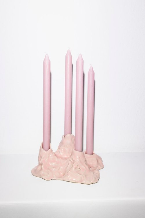 Ein rosa, handgeformter Kerzenständer aus glasiertem Ton mit vier eingesetzten rosafarbenen Stabkerzen, auf einem weissen Sockel vor einer weissen Wand.