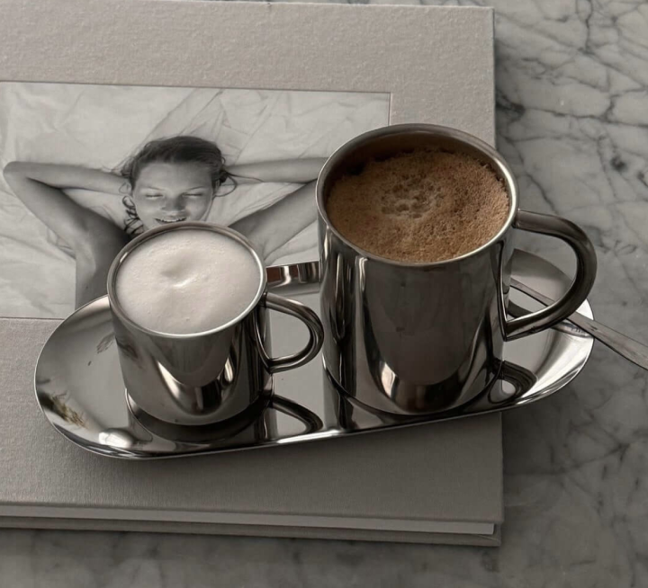Minimalistische Edelstahl-Tassen mit Cappuccino und Milchkaffee auf poliertem Edelstahl-Tablett, arrangiert auf einem Bildband auf Marmortisch