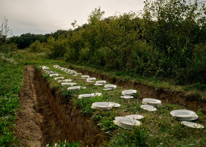 Langes, in die Erde eingelassenes Tischareal mit Tellern und eingewachsenem Grün inmitten eines offenen Feldes.