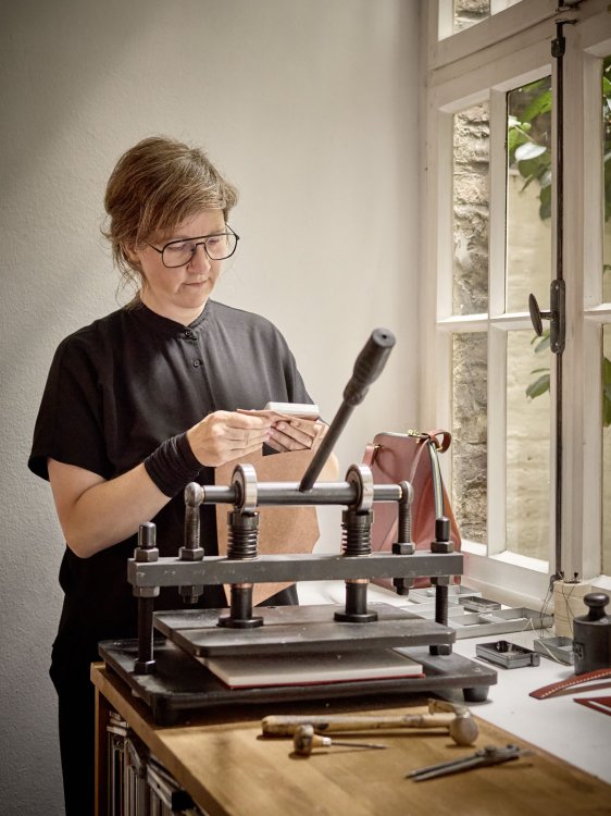 Designerin arbeitet in einem Atelier an einer Lederpresse, Tageslicht fällt durch ein Fenster auf den Arbeitstisch.