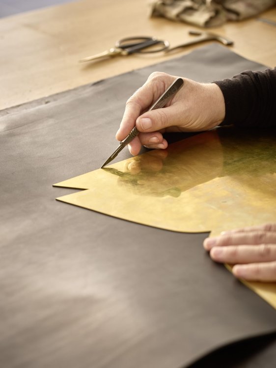 Hand schneidet mit einem Skalpell eine Form aus einer Schablone auf Leder, Werkzeuge liegen im Hintergrund.