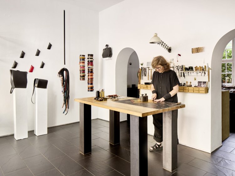 Grosszügiges Atelier mit Werkbank, Werkzeugwand und ausgestellten Ledertaschen, eine Person arbeitet am Tisch.