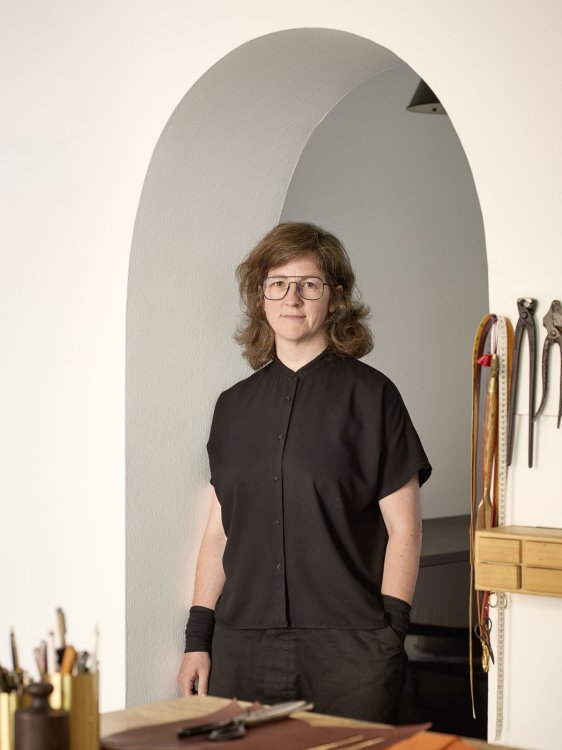 Designerin steht im Atelier vor einem Arbeitstisch, Werkzeuge und Materialien befinden sich im Hintergrund.