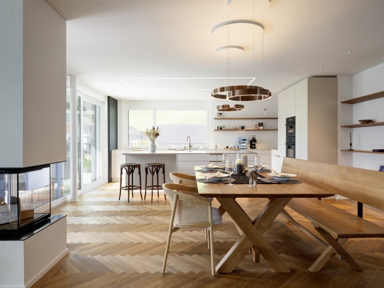 Blick in die Küche, links am Bildrand ein Cheminee, rechts ein Esstisch mit Stühlen, alles aus Holz. Im Hintergrund eine grosse, weisse Kücheninsel.