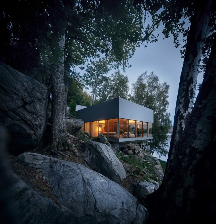 Modernes, kubisches Haus mit großen Glasfronten, das auf einem felsigen Hang zwischen Bäumen steht; warm beleuchteter Innenraum kontrastiert mit der kühlen Dämmerung, im Hintergrund ein See sichtbar.