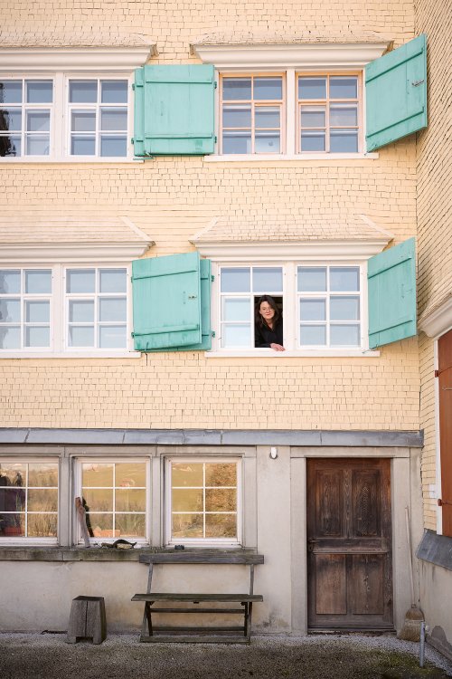 Gelbes Appenzellerhaus mit grünen Fensterläden; eine Frau lehnt aus einem geöffneten Fenster im oberen Stockwerk, darunter eine Holzbank und eine Tür im Erdgeschoss