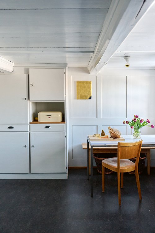 Heller, minimalistischer Küchen- und Essbereich mit weissen Einbauschränken, kleinem Tisch mit Stuhl, Blumenstrauss in Vase und Brot auf dem Tisch.