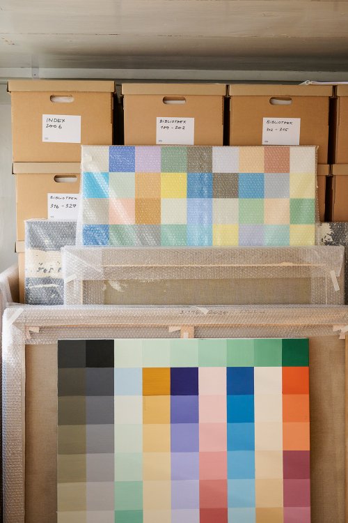Mehrere gerahmte Farbtafeln in Luftpolsterfolie lehnen vor gestapelten Archivkartons mit Beschriftungen in einem Lagerraum.