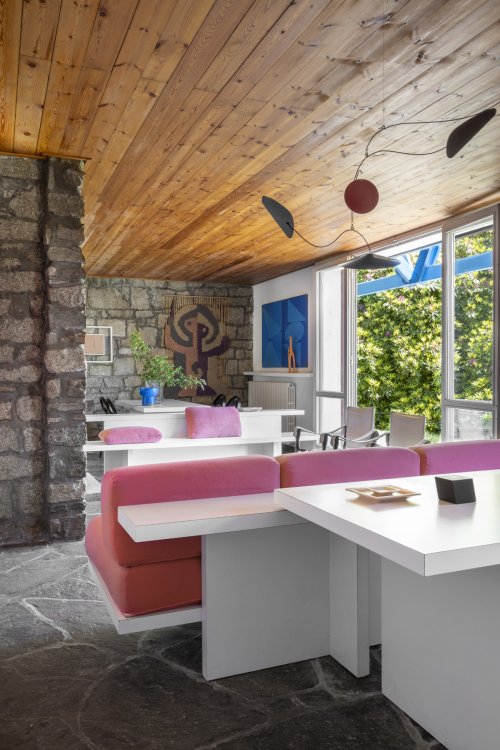 Blick ins Wohnzimmer mit massgefertigten weissen Einbaumöbeln und rosa Polster. Die Wände sind aus Stein, in der Mitte hängt ein Wandteppich mit braunem Muster.