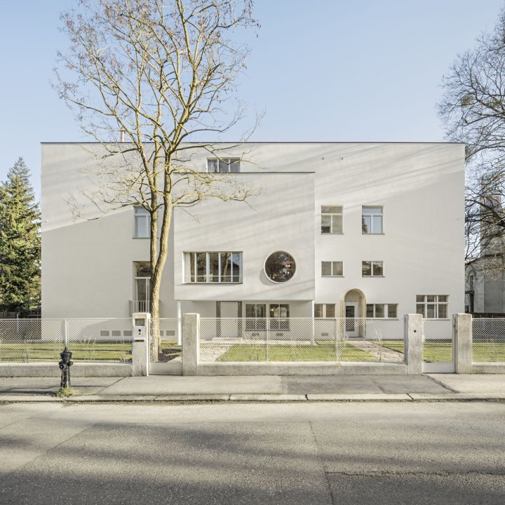 Ein weisse kubisches Gebäude mit Flachdach, davor eine grüne Rasenfläche und ein Maschendrahtzaun.