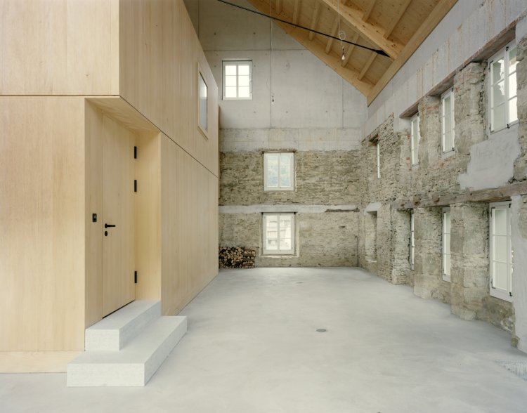 Ein altes Bauernhaus von Innen. Die bestehenden Mauern sind noch sichtbar und mit Beton gesichert. Im Inneren steht einen Holzkubus, der nachträglich als Wohnkörper integriert wurde.