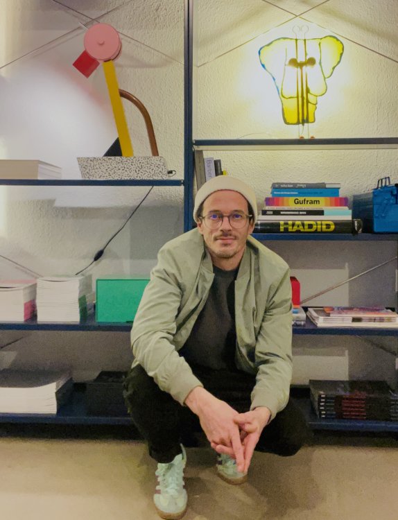 Demian Caviezel mit Mütze und Brille kauert vor einem Büchergestellt, in dem auch einige Objekte von Italien Radical Design stehen.