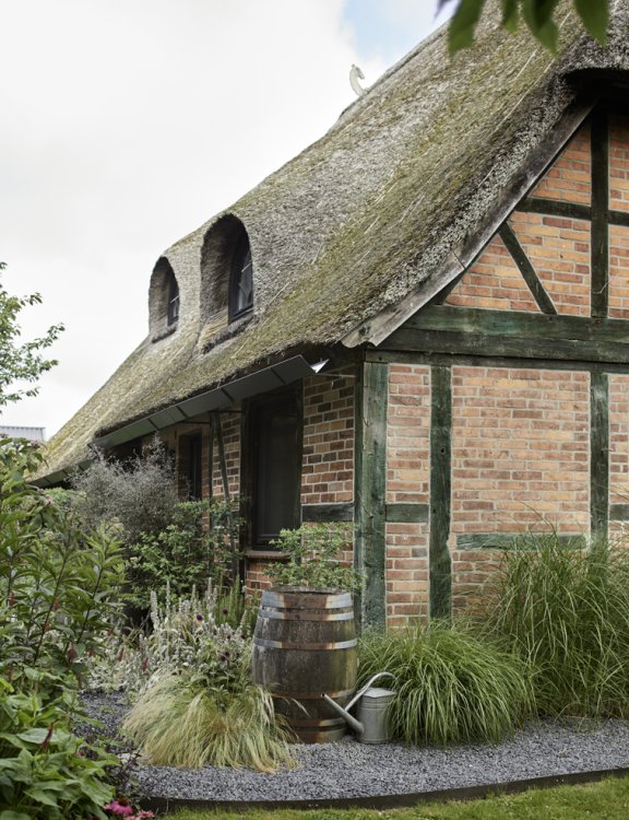 Eine Ecke eines Riegbaus aus roten Ziegeln und mit einem Reetdach. Davor sind Pflanzentöpfe und Gräser zu sehen.