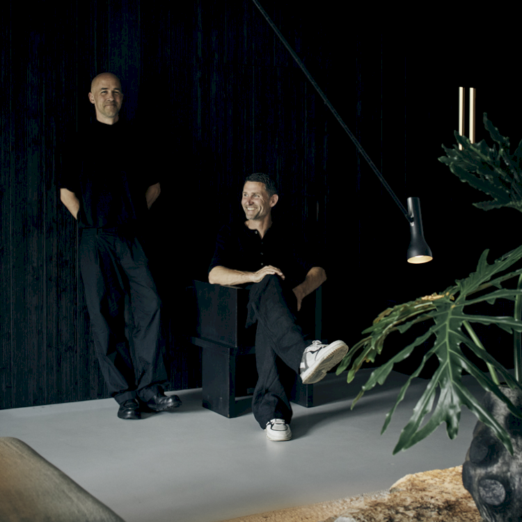 Porträt der beiden Floristen Moritz Mahl­stedt (links stehend) und Mario Leonhardt (rechts sitzend) in ihrem dunklen gehaltenen Wohnzimmer.