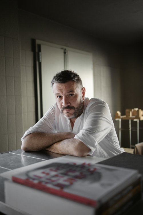 Der Architekt Lando Rossmaier sitzt in einem weissen Hemd an einem Tisch und blickt in die Kamera. Die verschränkten Arme sind auf der Tischplatte abgestützt.