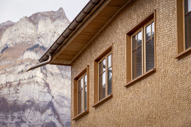 Ein Ausschnitt einer frisch sanierte Holzschindelfassade mit Holzfenstern direkt unter dem Dachabschluss. Im Hintergrund ragt eine Bergspitze gegen den Himmel empor.