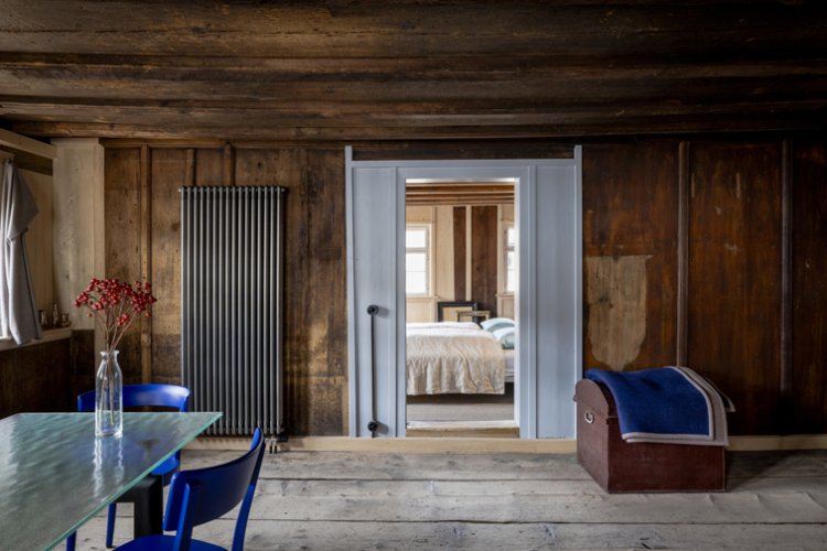 Eine Stube mit Holzboden, Holztäfer und tiefer, dunkler Holzdecke. Im Raum stehen ein Glastsich, blaue Holzstühle, eine Holztruhe mit blauer Wolldecke darauf. An der Wand steht ein fast raumhoher moderner Heizkörper. Der Blick geht durch einen weiss gestrichenen Türrahmen hindurch in ein gemütliches Schlafzimmer.