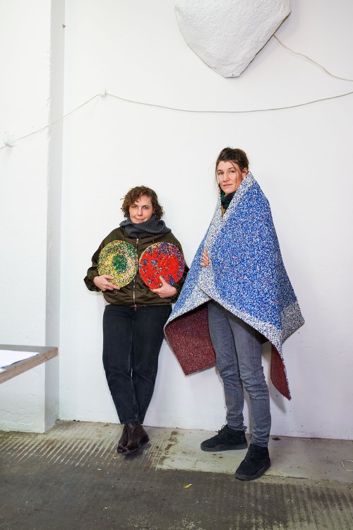 Die beiden Designerinnen Sarah Kueng und Lovis Caputo vom Duo Kueng Caputo stehen in ihrem Zürcher Atelier vor einer weissen Wand, Lovis Caputo hält zwei runde farbige Gegenstände in den Händen, Sarah Caputo hat einen blauen Teppich aus Gummigranulat über die Schultern gelegt.