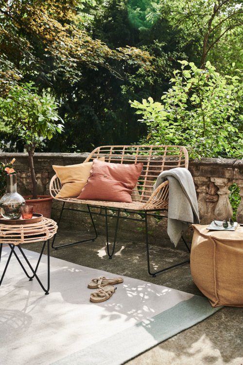 Eine Stein-Terrasse mit einer geflochtenen Sitzbank und passendem Beistelltisch mit bunten Kissen in rot und gelb, auf einem hellen Outdoorteppich, im Hintergrund sind grüne Bäume und Äste zu sehen.