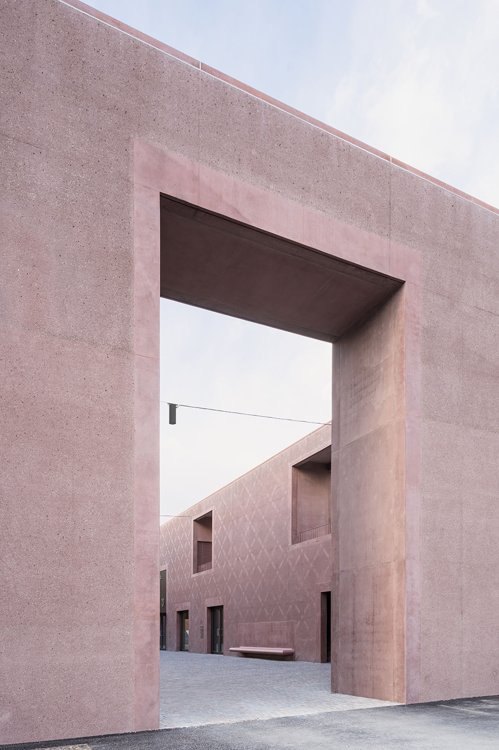 Blick durch ein grosses Eingangstor aus rötlich eingefärbtem Sichtbeton, das in den ummauerten Hof der neuen Musikschule Brixen führt.