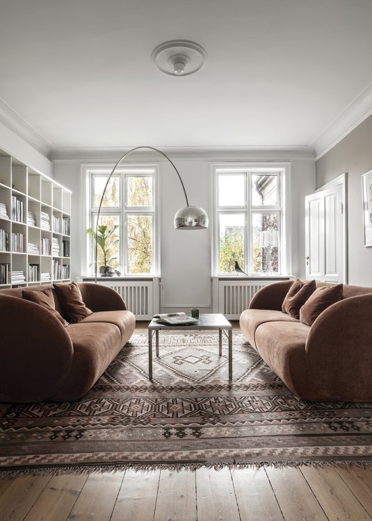 Ein Wohnzimmer in einem hellen Altbau mit Holzparkett, in dem zwei sich gegenüberstehende dunkelbraune, organisch geformte Sofas auf einem dunkelbraunen Kilim platziert sind. Links im Bild ist eine Bibliothek zu sehen.