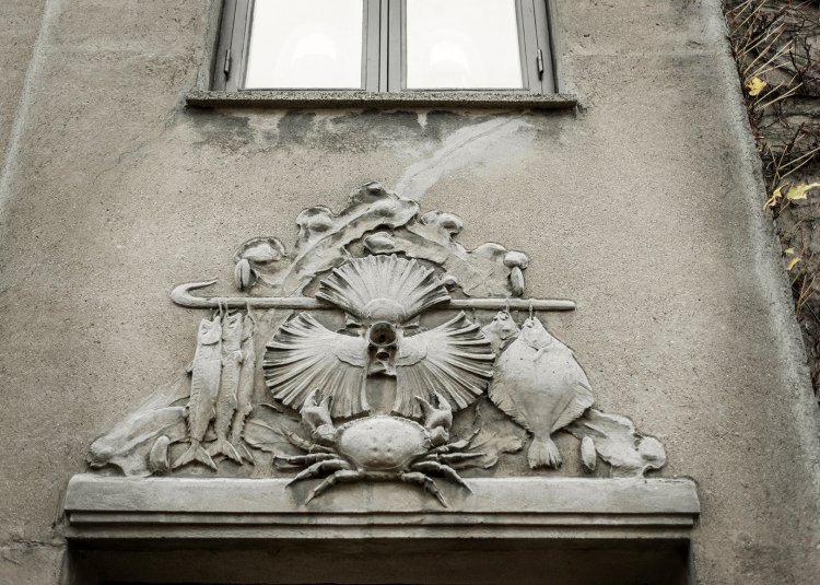 Ein detailreiches Steinrelief mit Fischen und Muscheln über einem Hauseingang mit grauer Fassade.