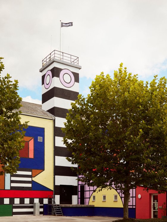 Aussenansicht der bunt gestalteten Montana-Fabrik in Haarby, Dänemark mit farbiger Fassade in geomtrischem Muster und einem schwarz-weiss-gestreiften Turm.