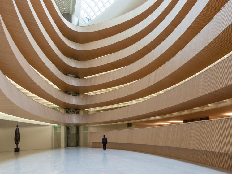 Innenansicht einer sechsgeschossigen, ovalen Halle mit Galerien. Ein grosses Glasdach überspannt den lichtdurchfluteten Raum.