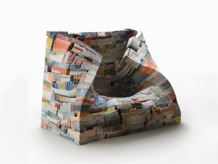 Ein sesselartiges Objekt aus unzähligen dünnen, bunt gefärbten Schichten, deren Kanten wie aufgeschlagene Buchseiten sichtbar bleiben, mit einer tiefen, unregelmässigen Mulde in der Mitte.