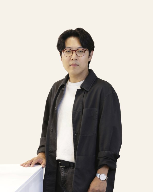 Porträt des Künstlers Jongjin Park: ein Mann mit dunklen Haaren, Brille und schwarzem Mantel über weissem T-Shirt, der vor neutralem Hintergrund steht, eine Hand auf einer weissen Fläche abgestützt.