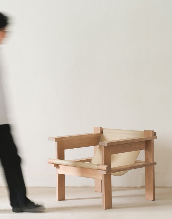 Ein Sessel aus hellem Holz mit locker gespannter, cremefarbener Ledersitzfläche steht im Raum, während eine Person daran vorbeigeht.